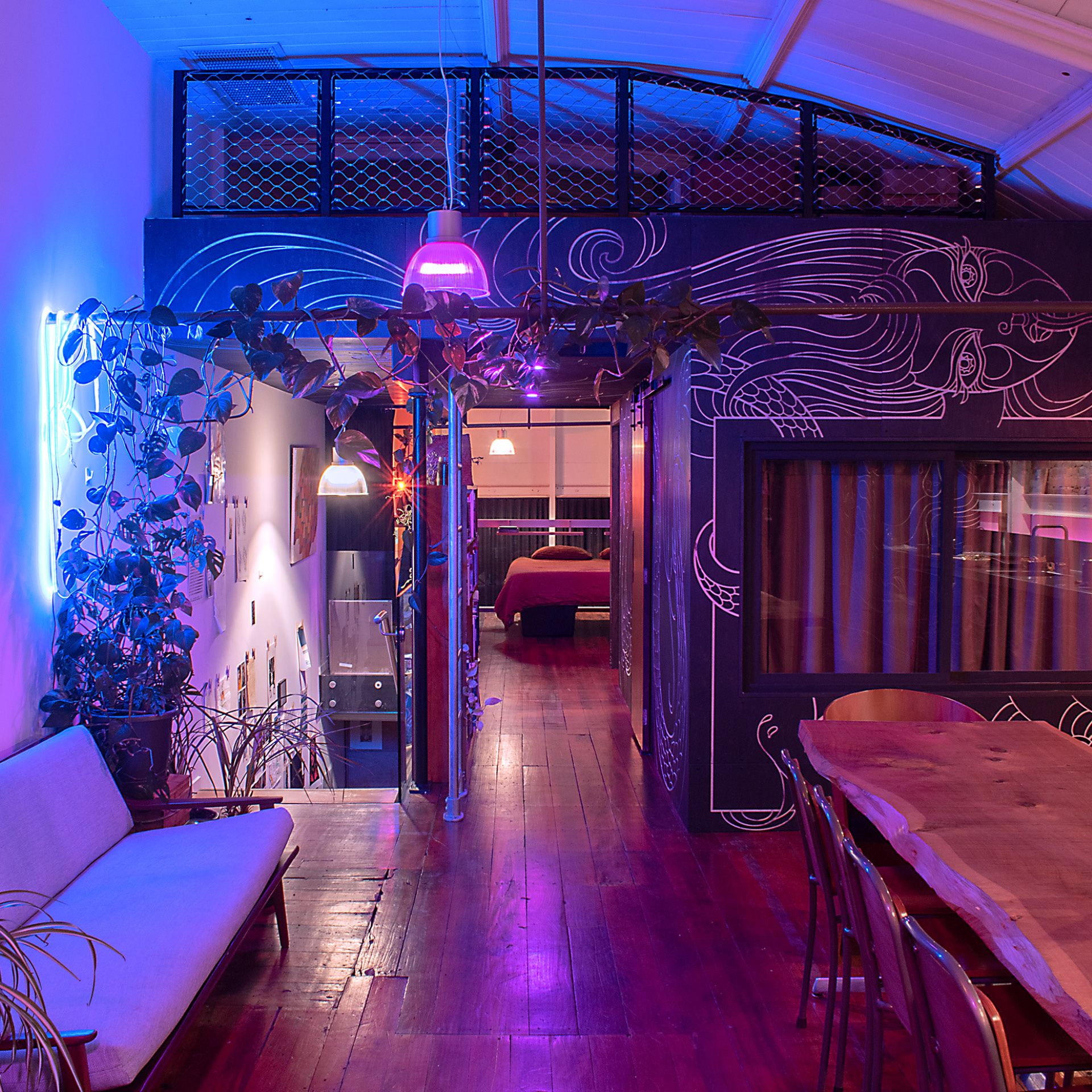 A beautiful room with plants, couches, and a mural above the door. The door in the centre opens into another room with a bed. The room is softly lit with blue and purple lighting.